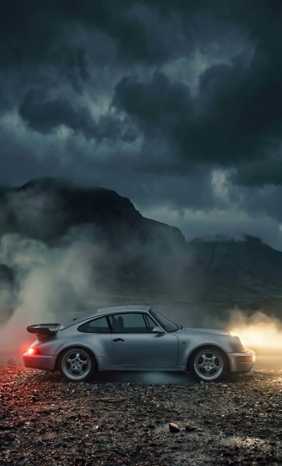 A silver sports car parked on dark gravel with mountains and heavy storm clouds behind it, mist drifting past