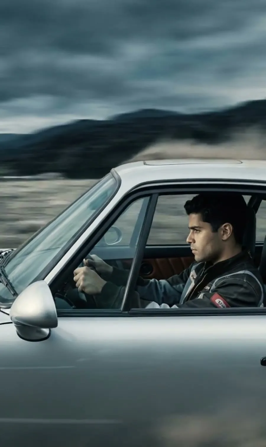 A young man driving a silver car, seen through the side window, the road and hills blurred with speed