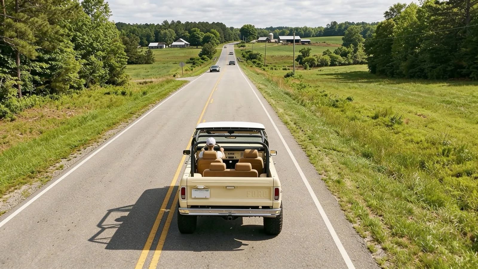 A pale yellow open-top truck seen from above and behind, driving down a two-lane country road between green fields