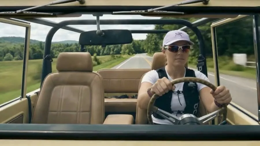 A woman in a cap and sunglasses at the wheel of an open-top truck with tan seats, a country road ahead