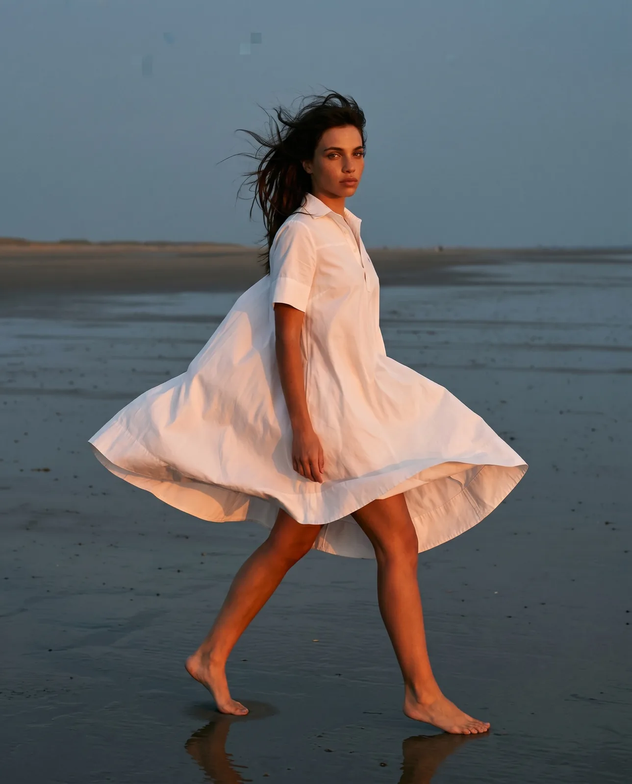 Woman in a white shirt dress striding barefoot across wet sand at dusk, the dress lifting in the wind