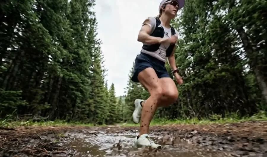 Runner in a cap, running vest and shorts splashing through a muddy puddle on a forest trail, seen from low down