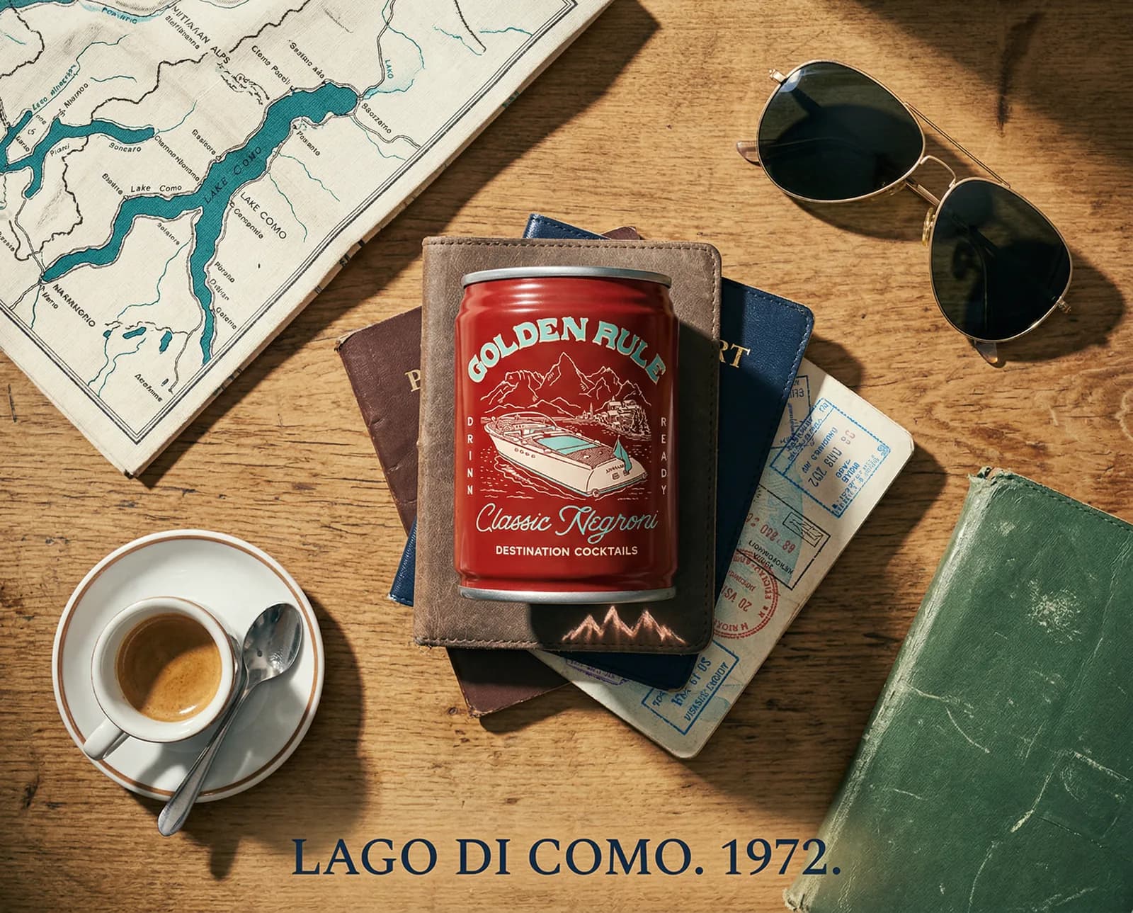 Flat lay on a wooden table, with a red can resting on a passport and a leather wallet, a folded map, an espresso cup and sunglasses around it